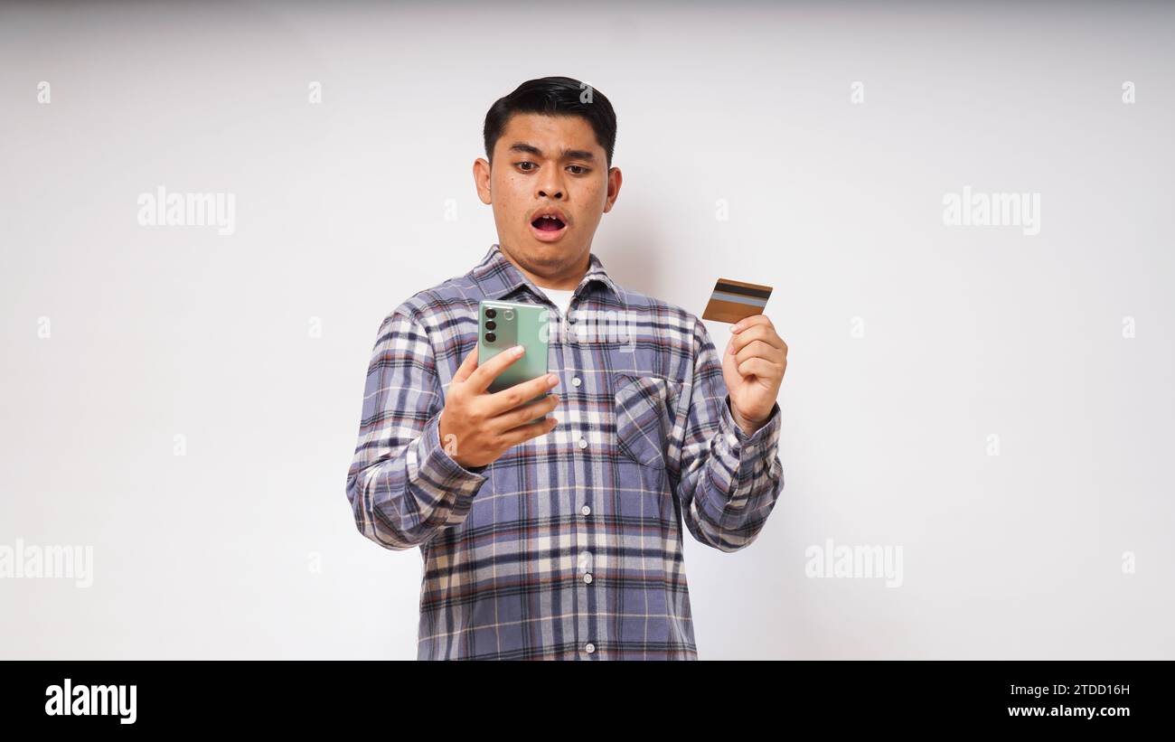 Asiatischer junger Mann mit schockiertem Ausdruck, der Kreditkarte und Smartphone hält, mit großen Augen auf den Smartphone-Bildschirm blickt, Innenstudio-Aufnahme isoliert Stockfoto