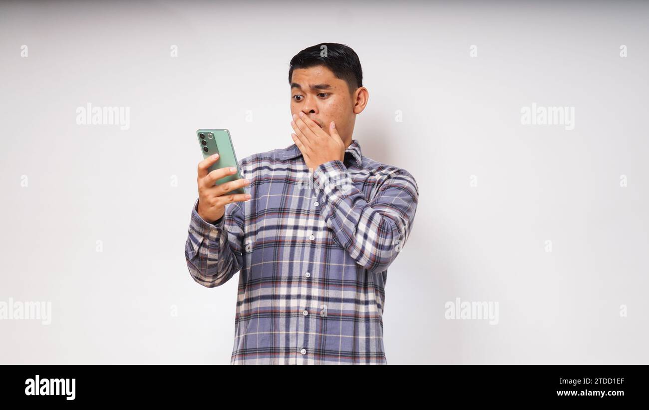 Junger asiatischer Mann mit schockiertem Gesichtsausdruck, während er ein Mobiltelefon auf weißem Hintergrund benutzt Stockfoto