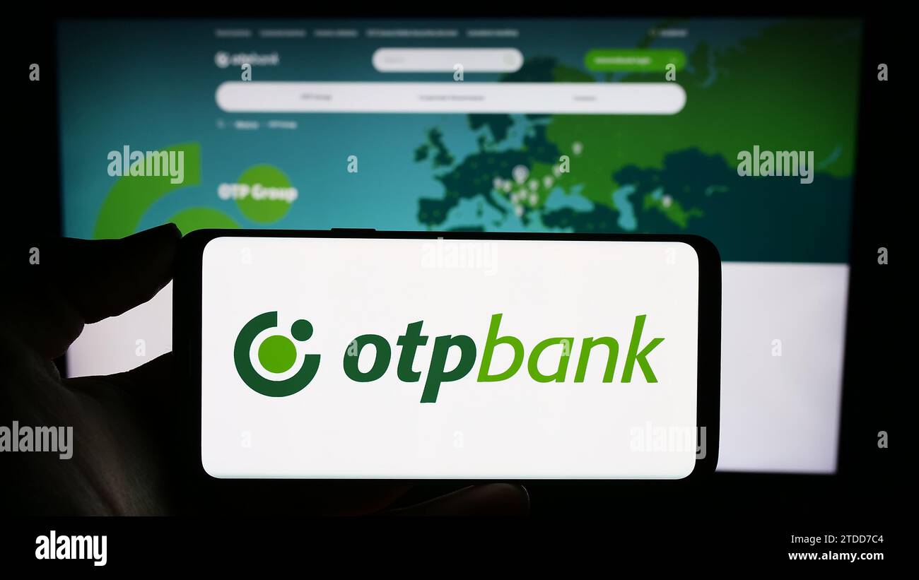 Person, die ein Mobiltelefon mit dem Logo des ungarischen Finanzdienstleisters OTP Bank Group vor der Website des Unternehmens hält. Schwerpunkt auf dem Display des Telefons. Stockfoto