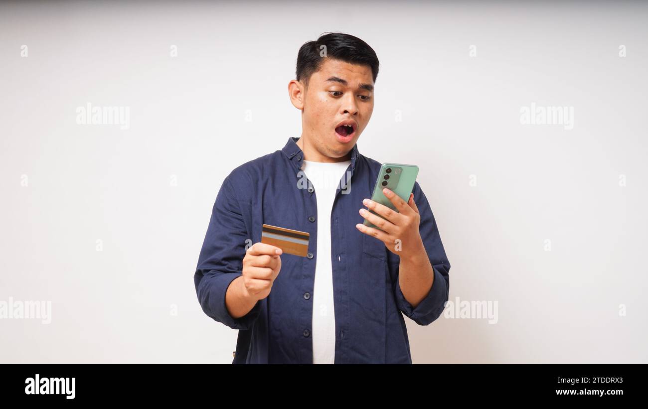 Asiatischer junger Mann mit schockiertem Ausdruck, der Kreditkarte und Smartphone hält, mit großen Augen auf den Smartphone-Bildschirm blickt, Innenstudio-Aufnahme isoliert Stockfoto
