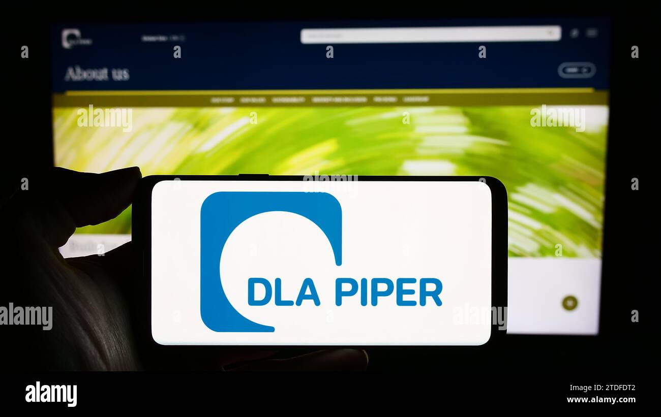 Person, die ein Mobiltelefon mit dem Logo der Anwaltskanzlei DLA Piper vor der Business-Webseite hält. Schwerpunkt auf dem Display des Telefons. Stockfoto