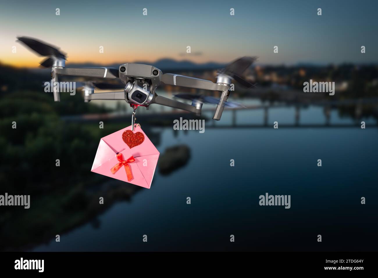 Drohne mit einem rosafarbenen Buchstaben mit rotem Herz und Bogen, der über einen See und eine Stadt im Hintergrund fliegt Stockfoto