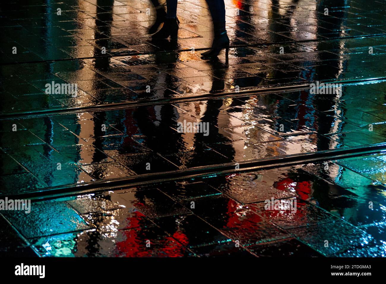 Die Silhouette der Fußgänger läuft in der Nacht im Regen entlang einer hell beleuchteten, schimmernden, nassen Straße. Stockfoto