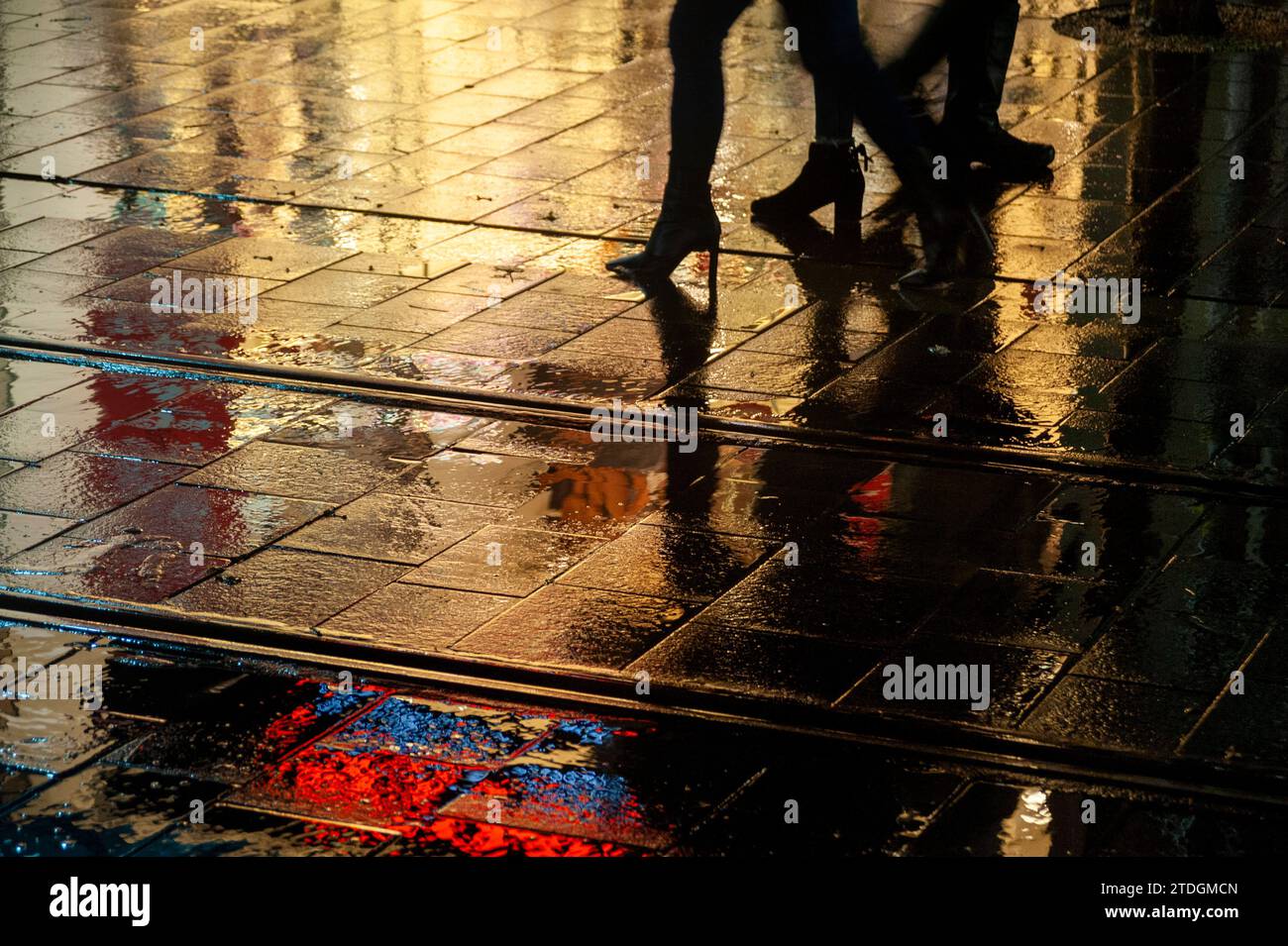 Die Silhouette der Fußgänger läuft in der Nacht im Regen entlang einer hell beleuchteten, schimmernden, nassen Straße. Stockfoto