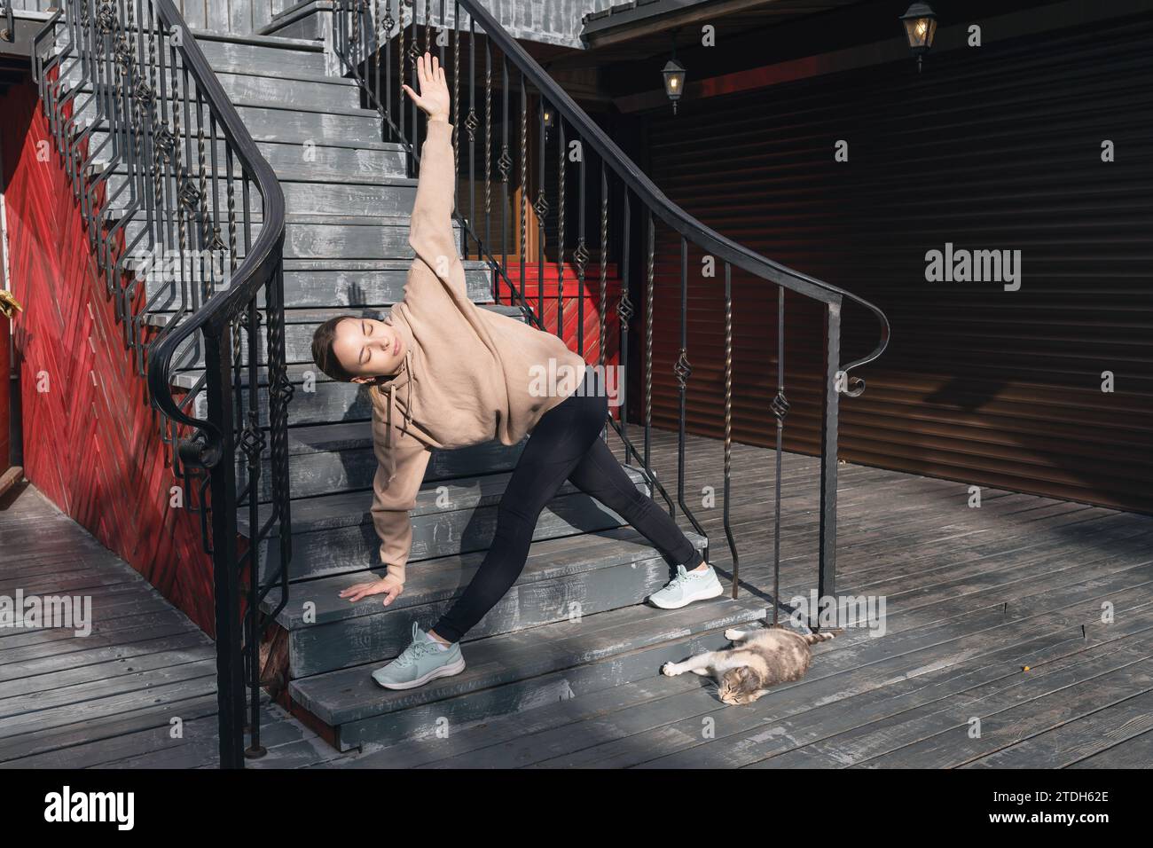 Eine junge Frau spielt auf der Straße in der Nähe einer Straßenkatze Sport, führt Trikonasana-Übungen, Dreiecksposen aus, übt Yoga in Sportbekleidung, die auf dem steht Stockfoto