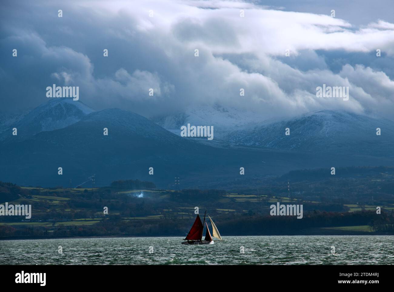 Ein traditioneller Ketch segelt auf den Menai Straights mit Snowdonia, der eine spektakuläre Kulisse bietet. Der Menai ist bekannt für seine extremen Gezeitenströme Stockfoto