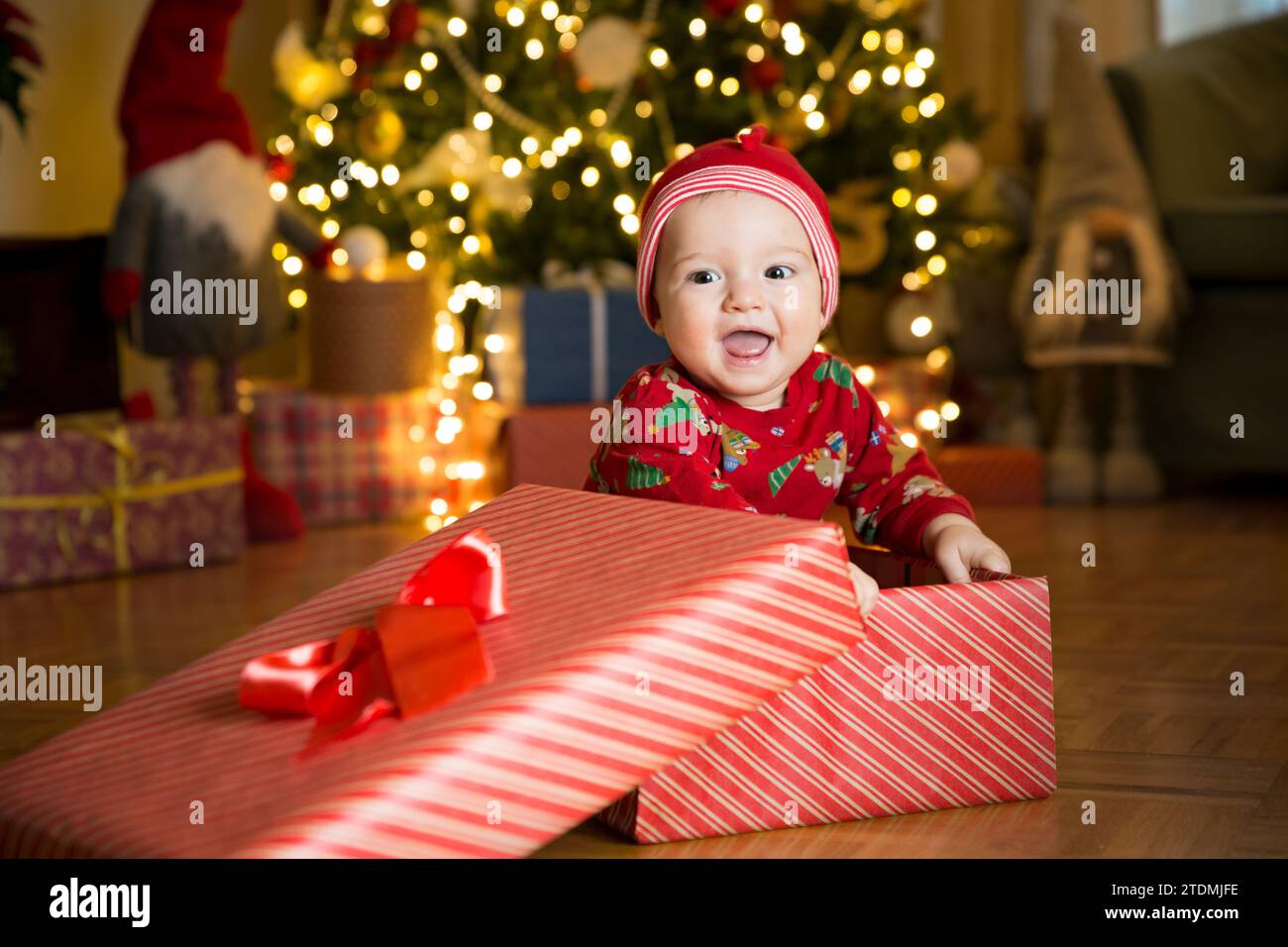 Glücklicher Junge in einer roten Mütze, der in einer Geschenkbox auf dem Hintergrund eines Weihnachtsbaums am Heiligabend sitzt. Frohe Feiertage, Neujahr. Gemütlicher, warmer Winterabend zu Hause. Weihnachtszeit Stockfoto