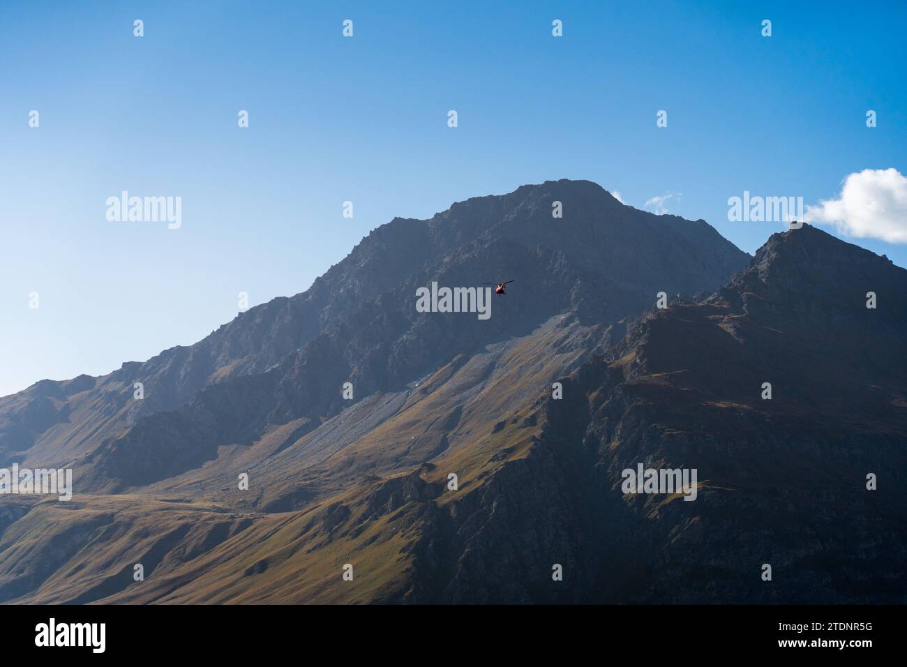 Roter Hubschrauber fliegt zum felsigen Berg Mont-Cenis für Rettungs- oder Sicherheitskontrollen. Blauer Himmel und sanftes Licht Stockfoto