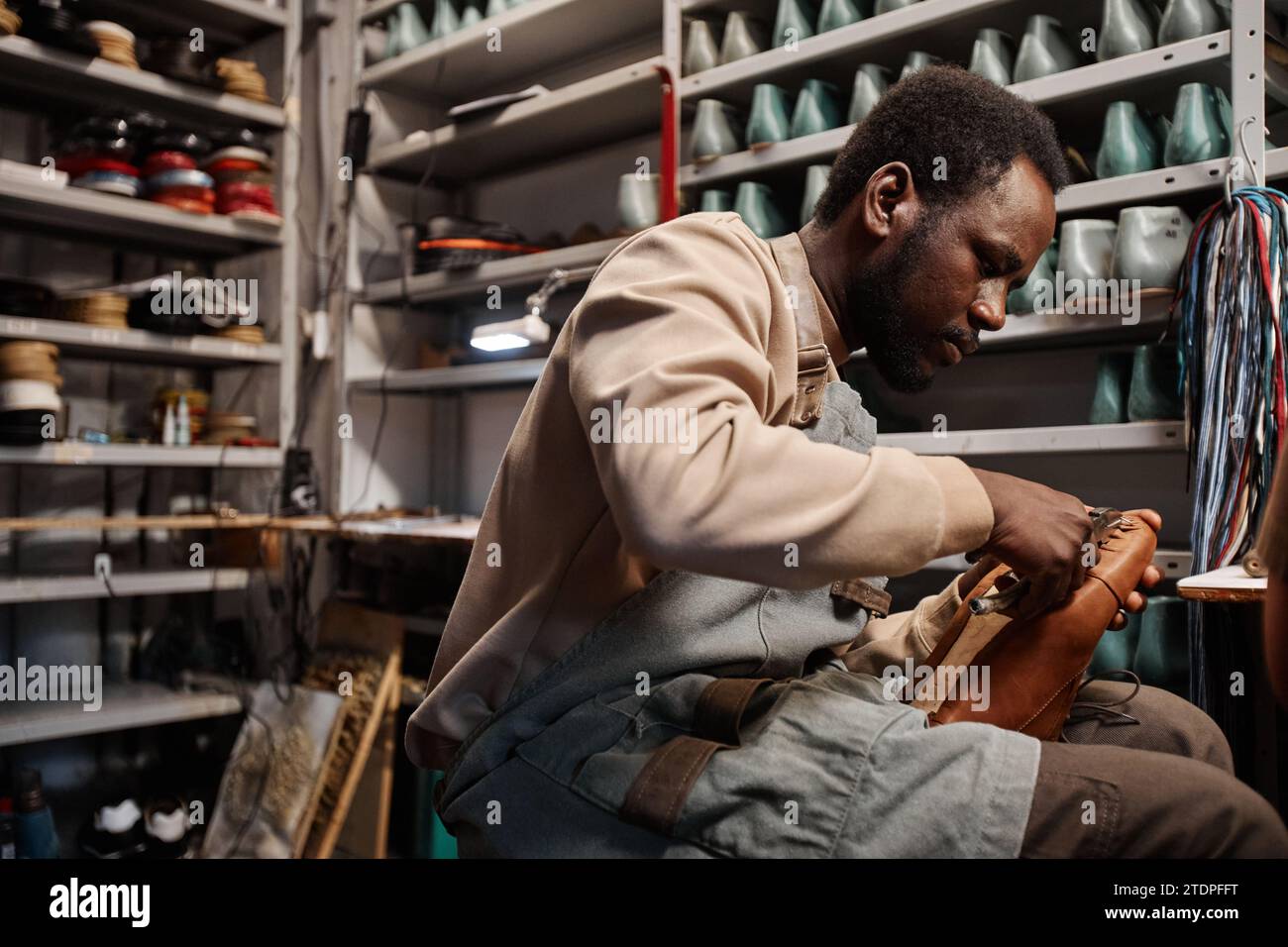 Junger afroamerikanischer männlicher Besitzer einer Schuhmacherei, der den oberen Teil des unfertigen Stiefels an der Sohle befestigt, während er am Arbeitsplatz sitzt Stockfoto