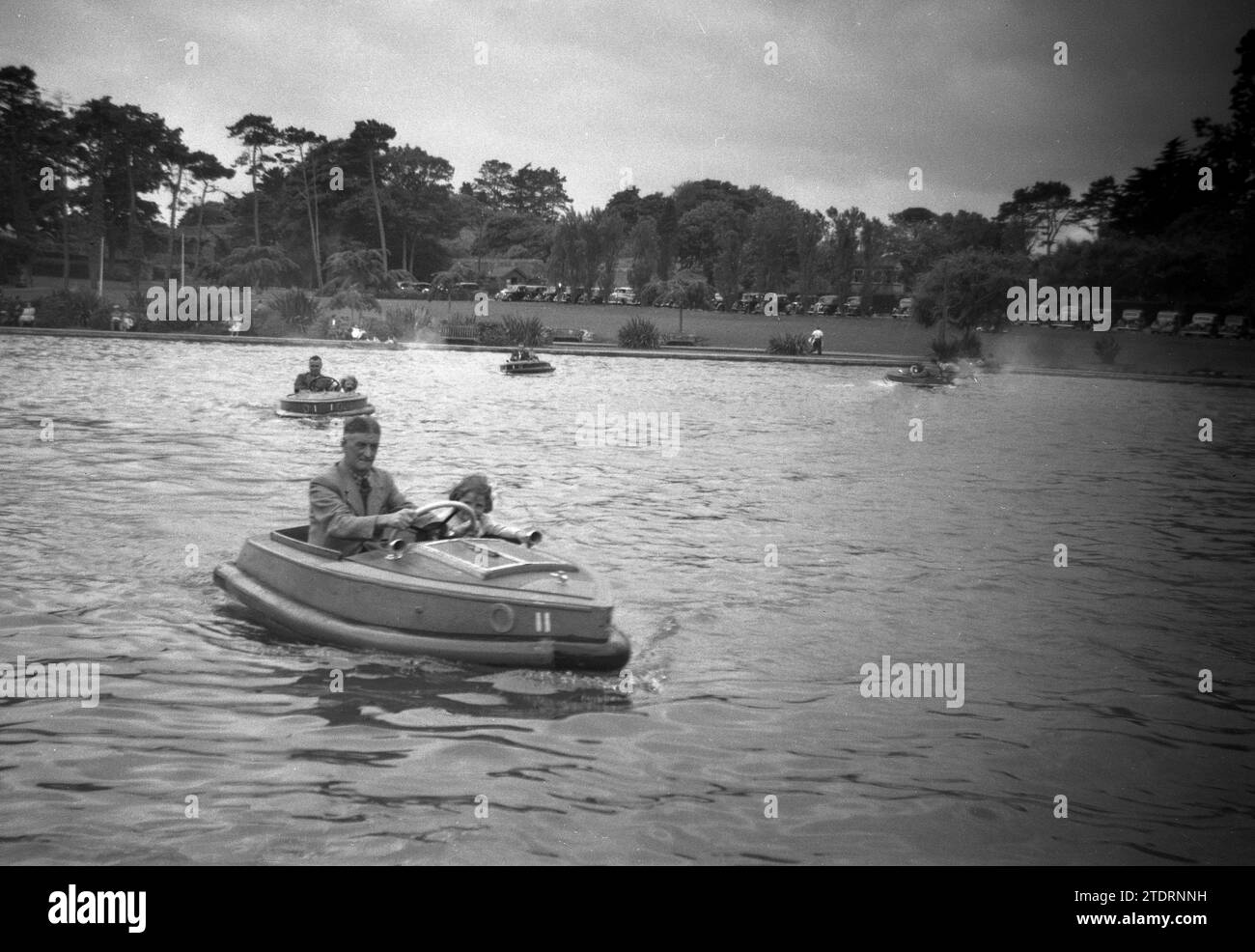 1950er Jahre, historisch, ein Großvater mit seiner Enkelin zusammen in einem kleinen zweisitzigen Motorboot mit Gummischutzverkleidung, auf dem Wasser eines großen Sees, England, Großbritannien. In der Ferne parkte eine Reihe von Autos aus der Zeit auf dem Gras neben dem See. Stockfoto