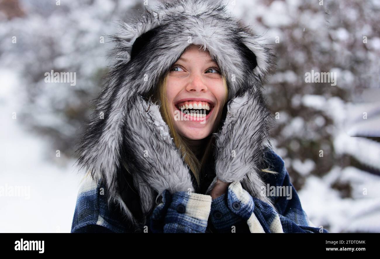 Winterzeit. Glückliche Frau, die den schneebedeckten Wintertag genießt. Kaltes Winterwetter. Lächelndes Mädchen in kariertem Mantel, Pelzmütze und Fäustlingen spazieren im Winterpark. Mode Stockfoto