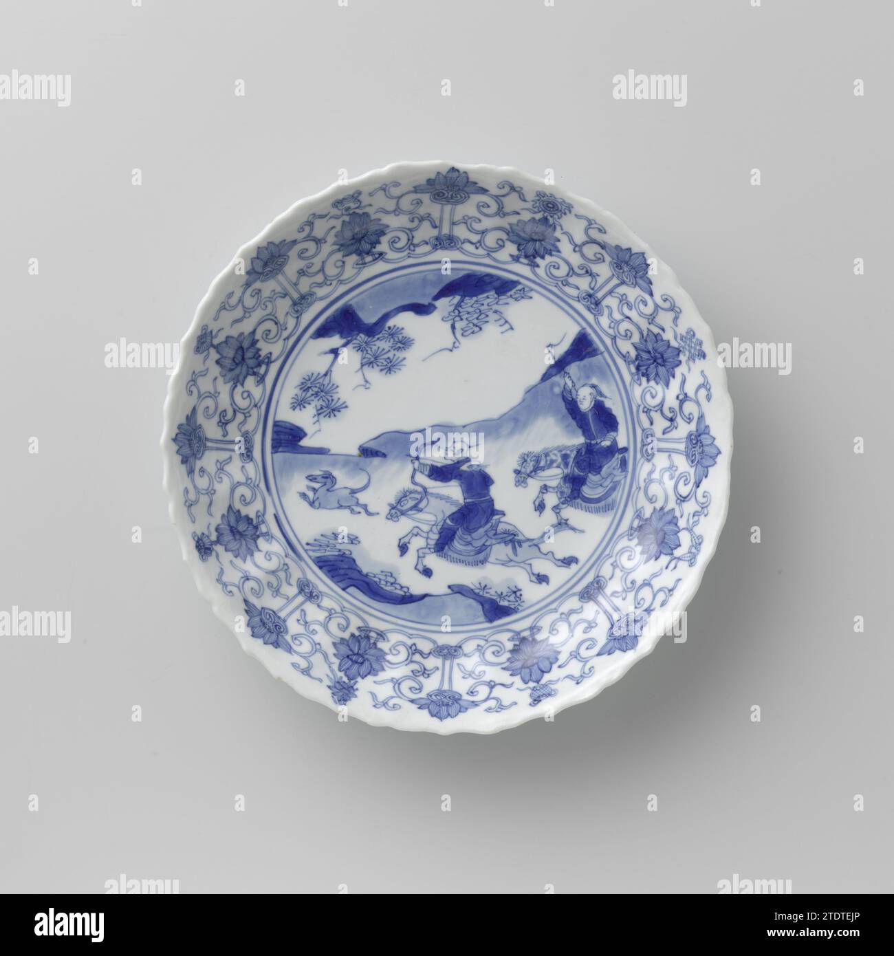 Untertasse mit Jagdszene, Lotusrollen und verheißungsvollen Symbolen, anonym, ca. 1700 - ca. 1724 Porzellanschale mit runder, geriffelter Wand und gewelltem Rand, in Unterlazblau bemalt. Auf der Wohnung eine Jagdszene mit zwei bewaffneten Reitern in einer Landschaft, die einen Hasen jagen; der Rand mit einem verbundenen Band von Lotusranken mit Ruyi-Motiv, zwischen dem die acht Glückssymbole (Chakra, Muschel, Sonnenschirm, Baldachin, Lotus, Vase, Angeln, unendlicher Knoten); der Rücken mit sechzehn losen Blütenzweigen. Auf der Unterseite mit einer nicht identifizierten sechsstelligen Marke in einem Doppelkreis gekennzeichnet. Einige Späne in der Kante. Blaue Wh Stockfoto