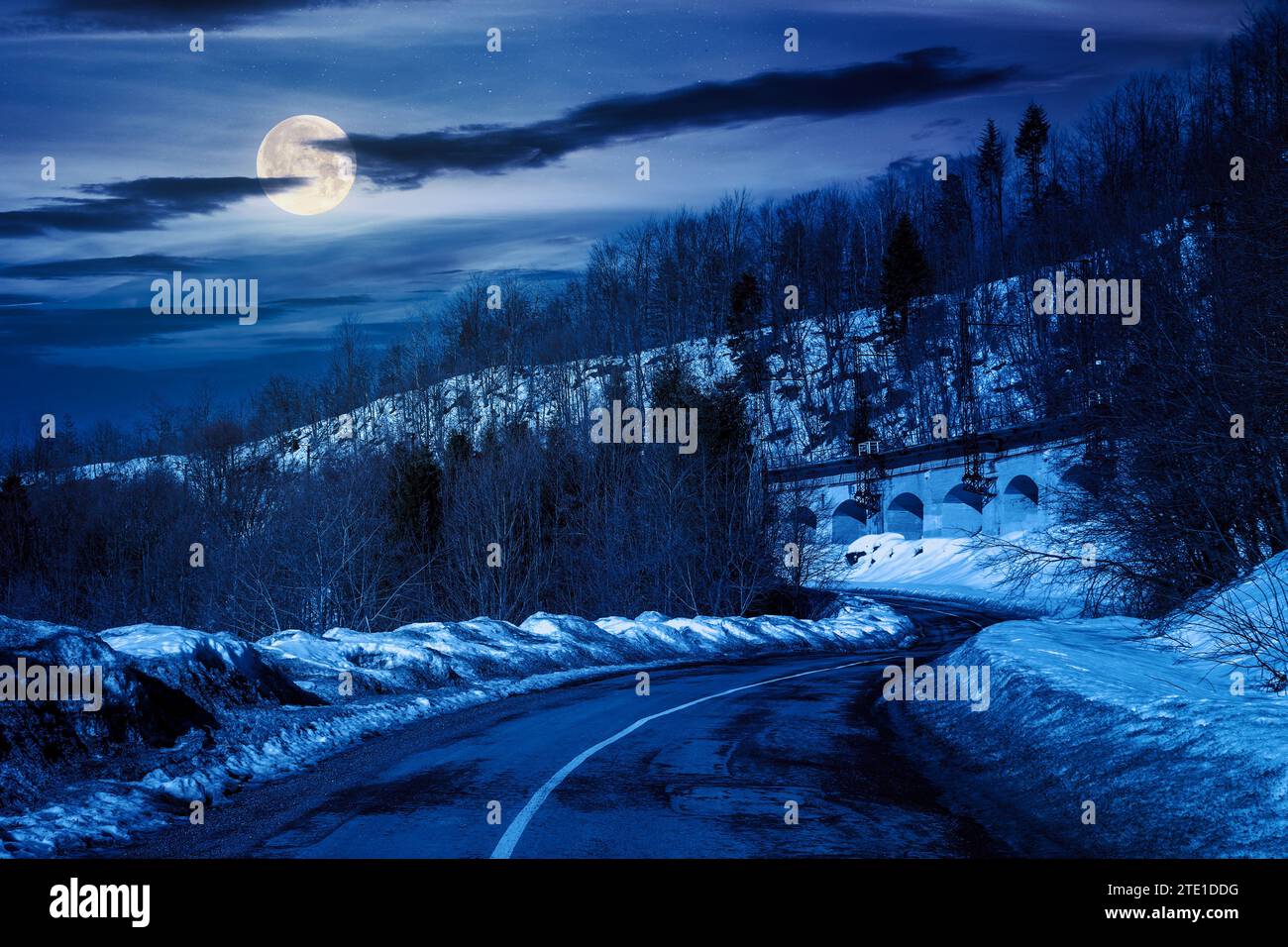 Der alte Straßenpass schlängelt sich nachts durch schneebedeckte Hügel. Bergige Winterlandschaft unter einem Himmel mit Wolken im Vollmondlicht Stockfoto