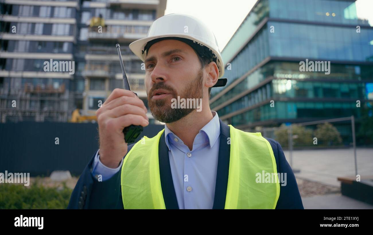 Moving Shot kaukasischer Baumeister in Helm und Sicherheitsweste spricht im Außenbereich im Außenbereich im Stadtbauarchitekt Bauingenieur Bauunternehmer Stockfoto
