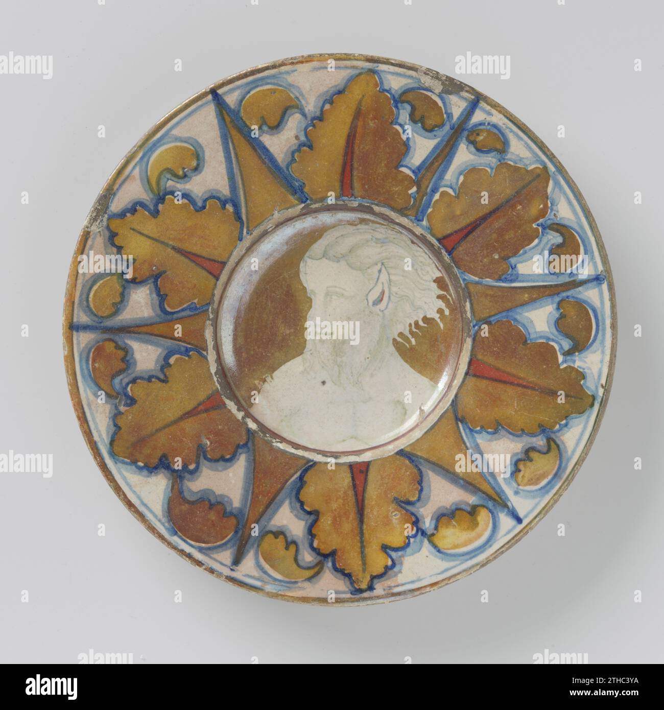 Platte mit vertiefter flacher, mehrfarbiger, mit Satyr bemalter, spitzer Ohren und Bart, anonym, ca. 1520 - ca. 1530 runde Platte aus mehrfarbig bemalter Majolika, mit vertiefter flacher Platte. Auf der Wohnung ist das linke Porträt des Mannes (Satyr) gemalt. Der Mann hat spitze Ohren und einen Bart. Am Rand waren Eichenblätter auffällig, durchsetzt von spitzen Formen. Die Platte ist in blau, Gold, braun und rot lackiert. Die Platine ist markiert. Gubbio Steingut. Zinnglasur. Bleiglasur Majolika Runde Platte aus mehrfarbig lackiertem Majolika, mit einer vertieften flachen Platte. Auf der Wohnung, das Porträt des Mannes (Satyr Stockfoto