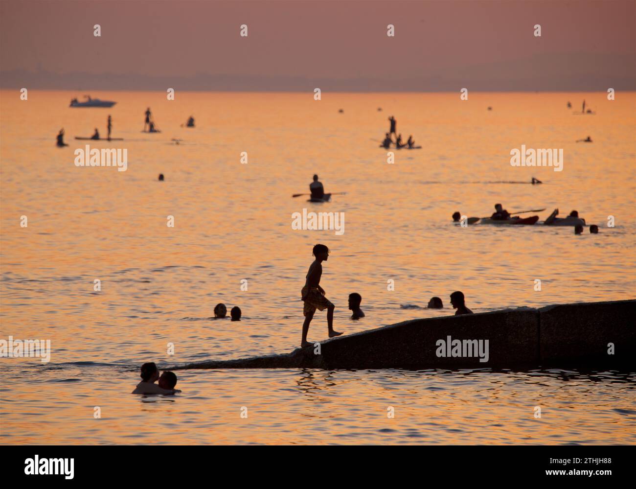 Silhouetten von Menschen, die den Strand bei Sonnenuntergang, Brighton & Hove genießen. Der Himmel erleuchtet mit dem schwindenden Licht des Tages. Boot- und Paddelboarder auf dem Meer in der Ferne. England, Großbritannien Stockfoto