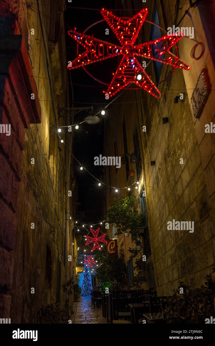 Rote, achtzackige maltesische Kreuzsterne hängen über einer engen Gasse in Birgu, Malta, mit einem beleuchteten Weihnachtsbaum am anderen Ende. Das Malteser Kreuz ist das traditionelle Emblem des Ordens von St. Johann, der Malteserritter. Stockfoto