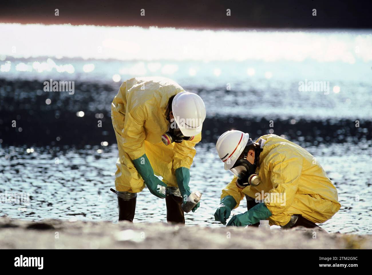 Zwei Techniker, die Schutzkleidung tragen, untersuchen giftige Abfälle, die an einem Flussufer gefunden wurden Stockfoto