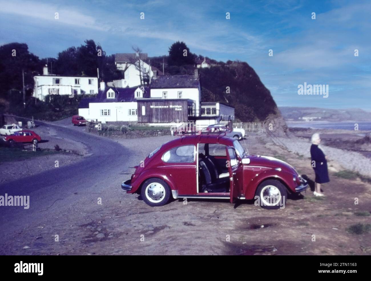 Ein rubinroter Volkswagen-Käfer-Wagen. In diesem Bild aus den 1970er Jahren stand eine Frau neben ihrem Volkswagen Käfer neben der Wisemans Bridge in Saundersfoot, Pembrokeshire in Wales, Großbritannien. Stockfoto