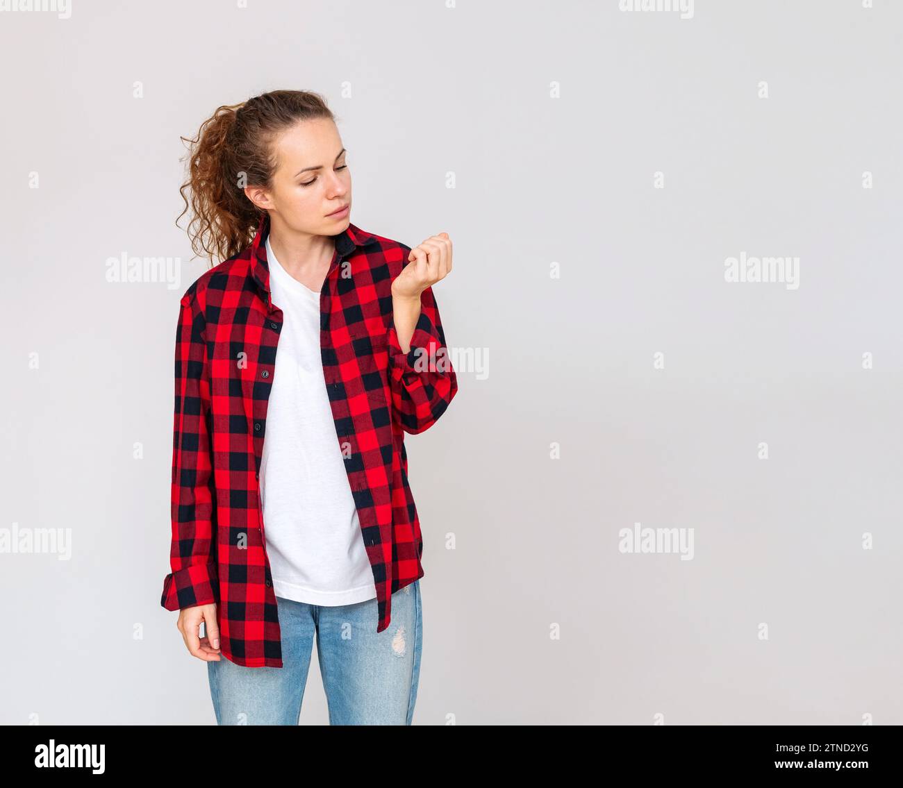 Lässig tragende Frau, die vor schlichtem Hintergrund steht und auf ihre Nägel blickt. Nicht gut gepflegte Hände. Stockfoto