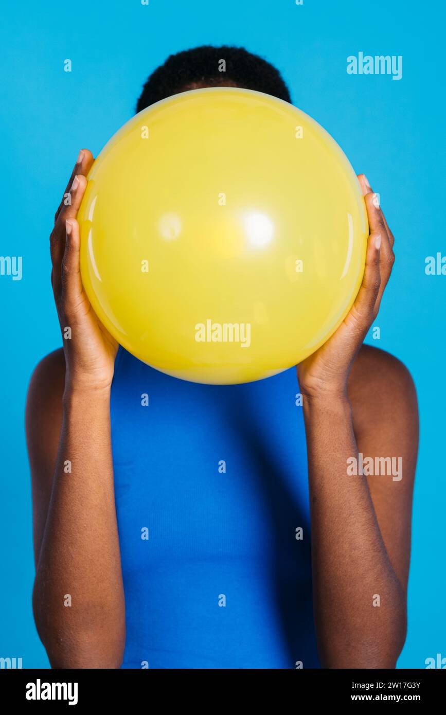 Frau, die einen gelben Ballon vor blauem Hintergrund in einem Studio bläst. Frau mit kurzen Haaren, die einen gelben Ballon aufbläht. Stockfoto