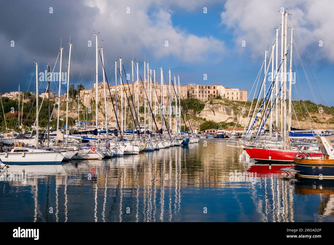 Segelboote vor Anker im Hafen und Jachthafen von Tropea, den Häusern der Stadt auf einem Felsen in der Ferne. Stockfoto