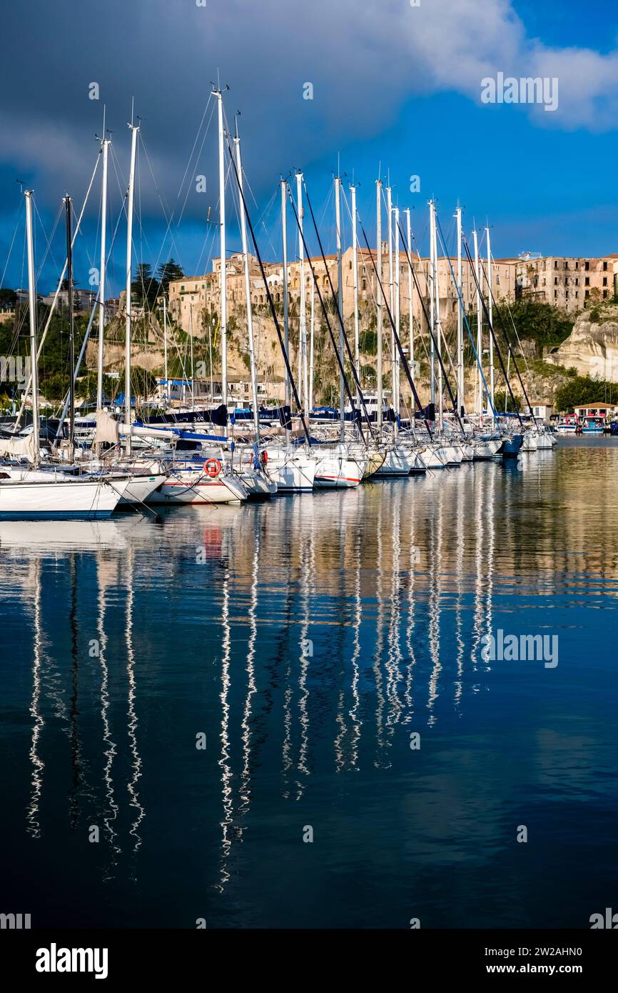 Segelboote vor Anker im Hafen und Jachthafen von Tropea, den Häusern der Stadt auf einem Felsen in der Ferne. Stockfoto