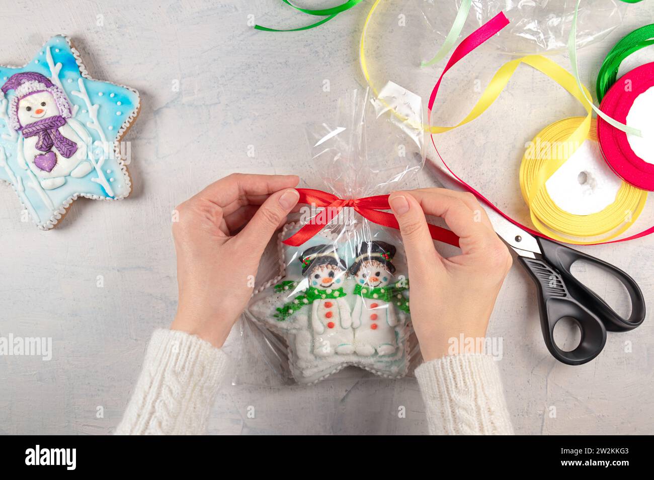 Weibliche Hände in einem weißen Pullover binden eine rote Schleife auf einem Paket Lebkuchenkekse mit Schneemännern auf dem Hintergrund eines Lichttisches, mehrfarbige Bänder und Scheren. Konzept für selbstgemachte Weihnachts- oder Neujahrsgeschenke. Stockfoto
