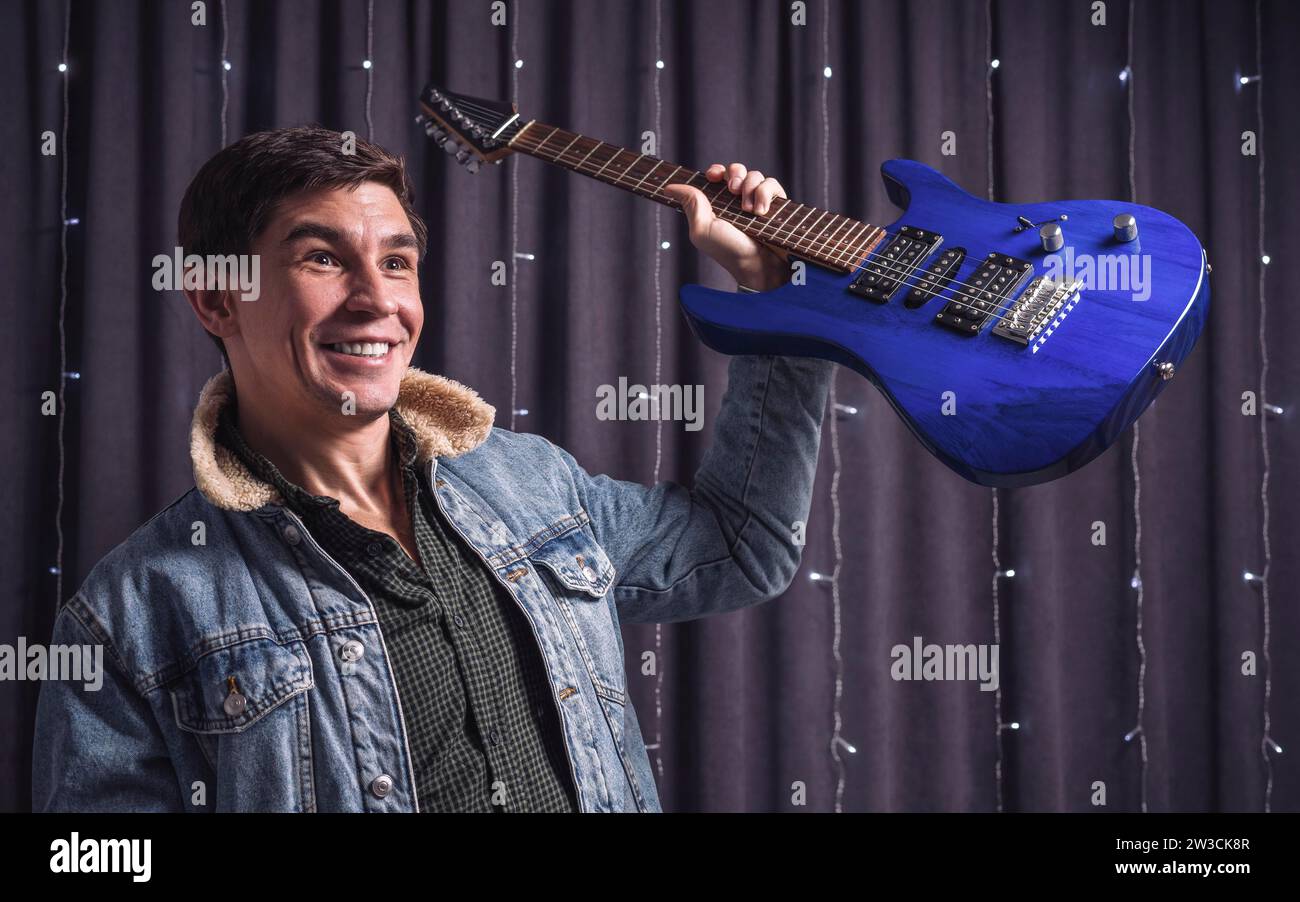 Bild eines emotionalen Musikers in einer Jeansjacke, der triumphierend eine blaue E-Gitarre hochhebt. Konzertkonzept. Gemischte Medien Stockfoto