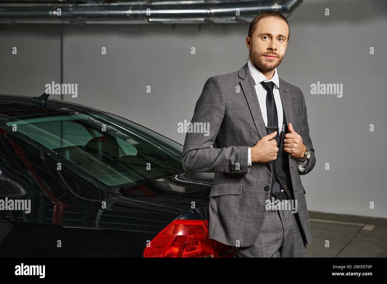 Attraktiver Mann im schicken Anzug, der in die Kamera schaut und vor seinem Auto posiert, Geschäftskonzept Stockfoto