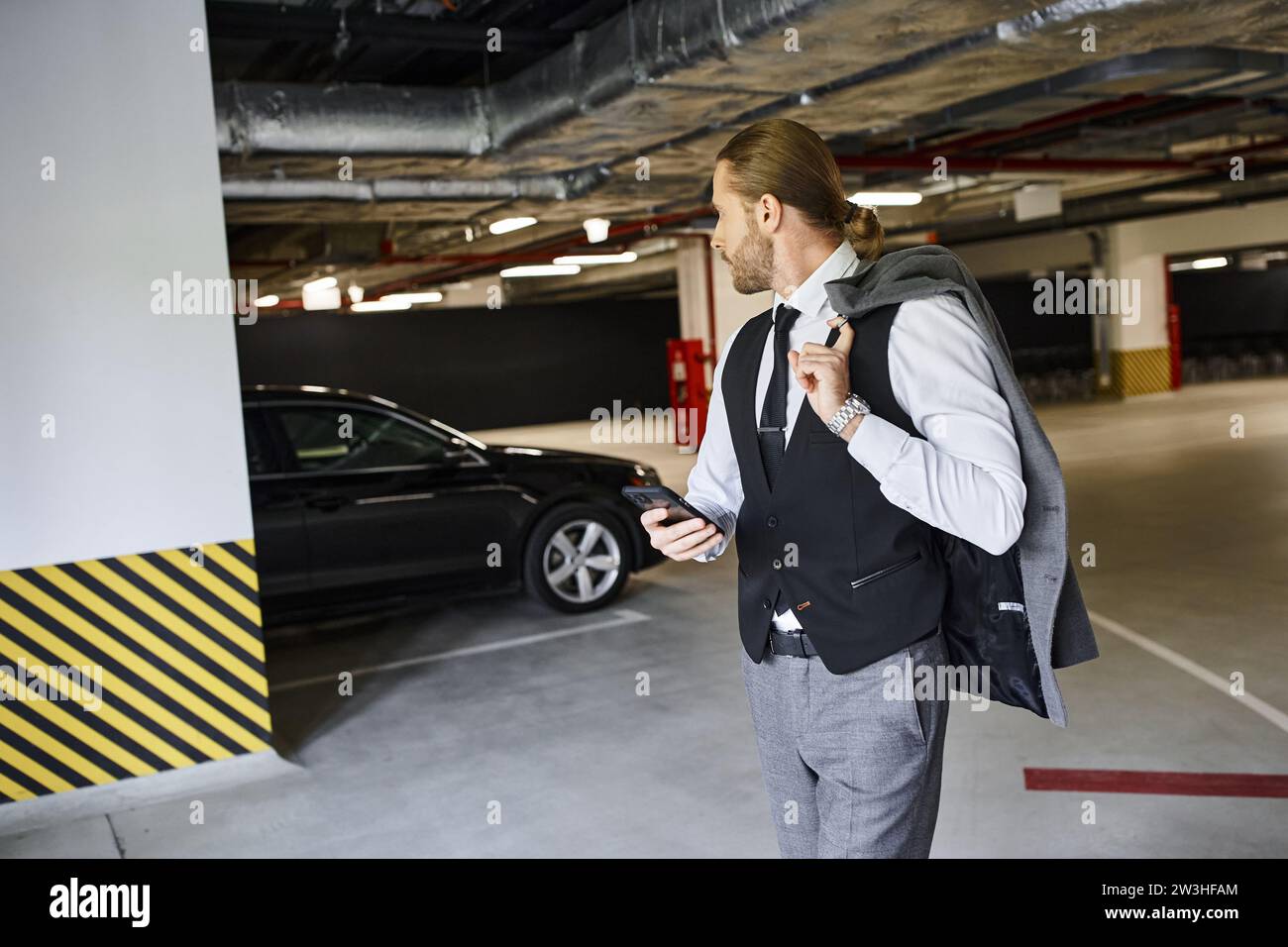 Gut aussehender bärtiger Mann in schwarzer Weste mit Handy und Blick auf sein Auto, Geschäftskonzept Stockfoto