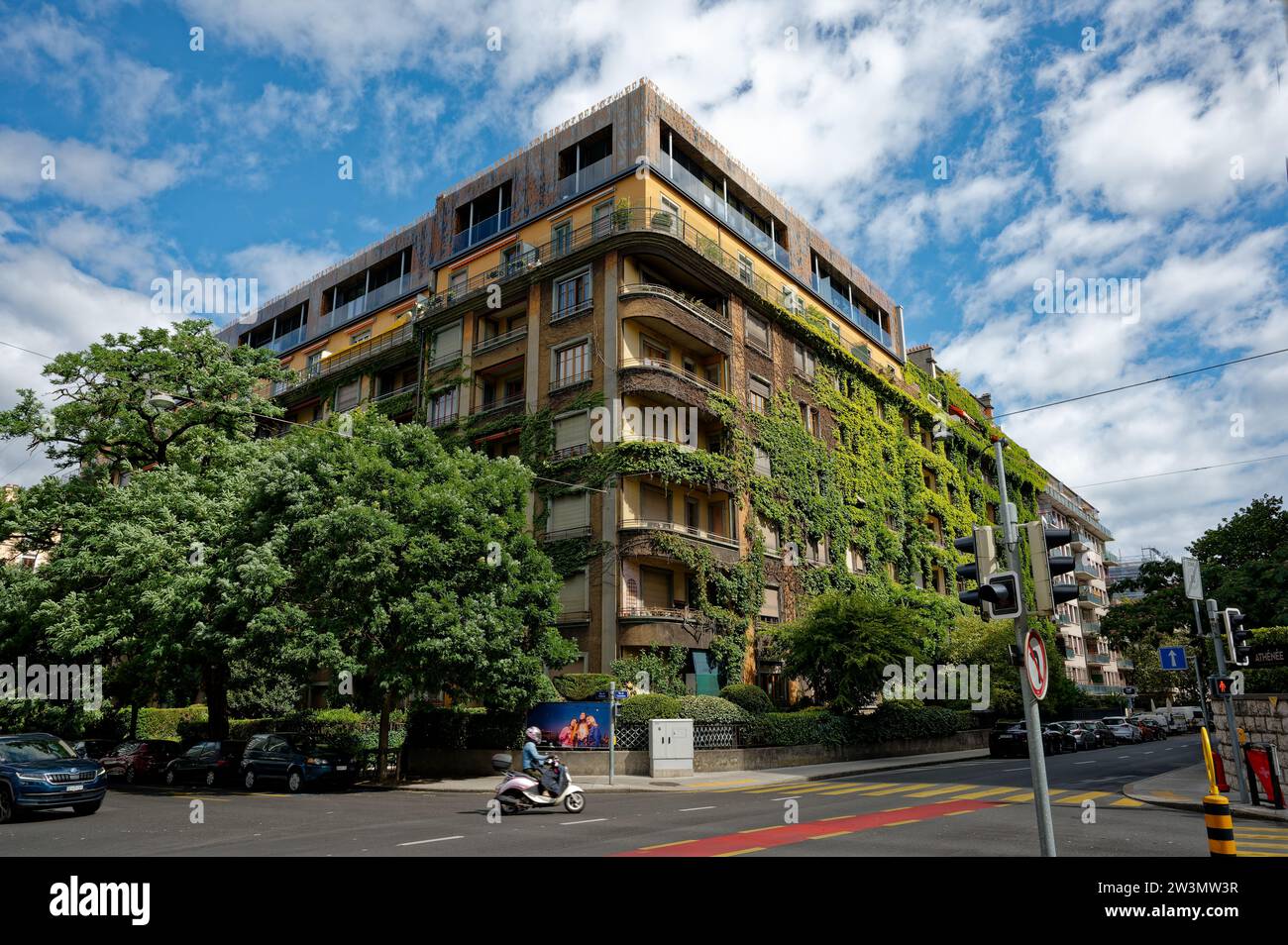 Ein imposantes Wohngebäude mit üppigem, hellgrünem Laub an der Außenseite, eingebettet in eine charmante Ecke von Genf, während ein Roller vorbeifährt. Stockfoto