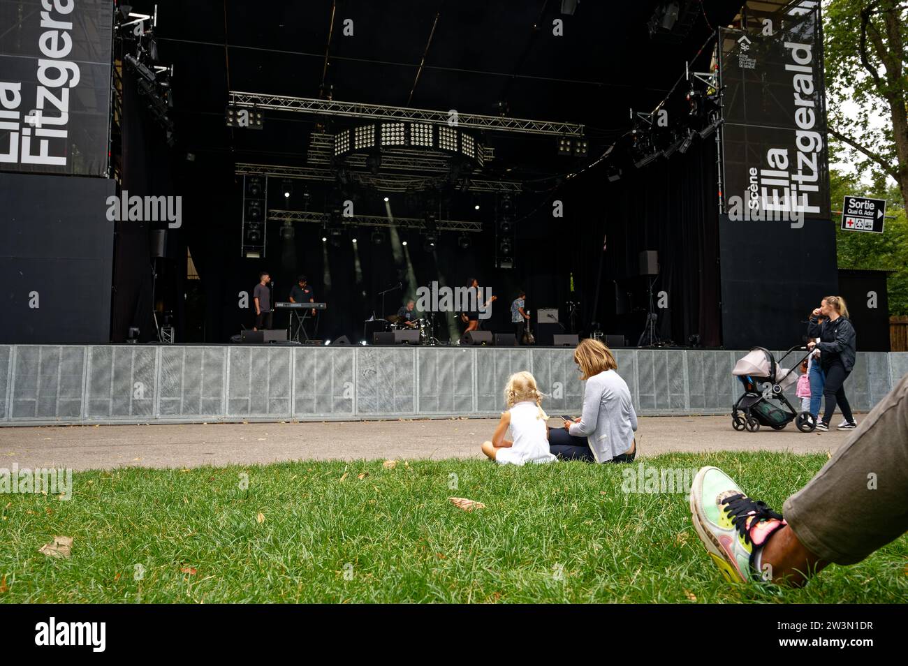 Die blonde Frau und ihre kleine Tochter entspannen sich auf dem üppigen Gras, ihre Aufmerksamkeit fesselt die talentierte Künstlerin Evita Koné und ihre dynamische Band. Stockfoto