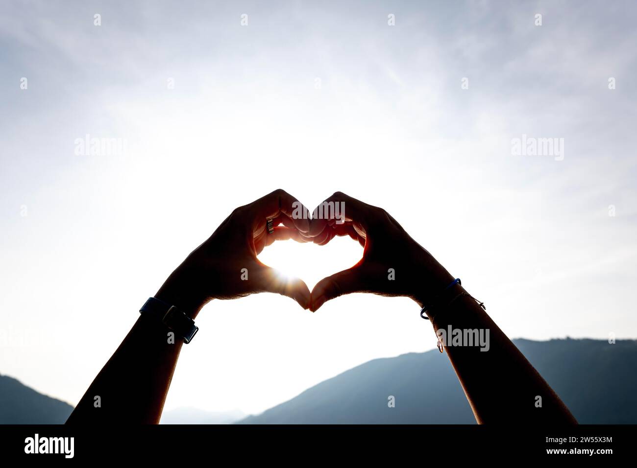 Frauenhände machen eine Herzform gegen den Himmel mit Sonne und Sonnenstrahl und Berg in der Schweiz Stockfoto