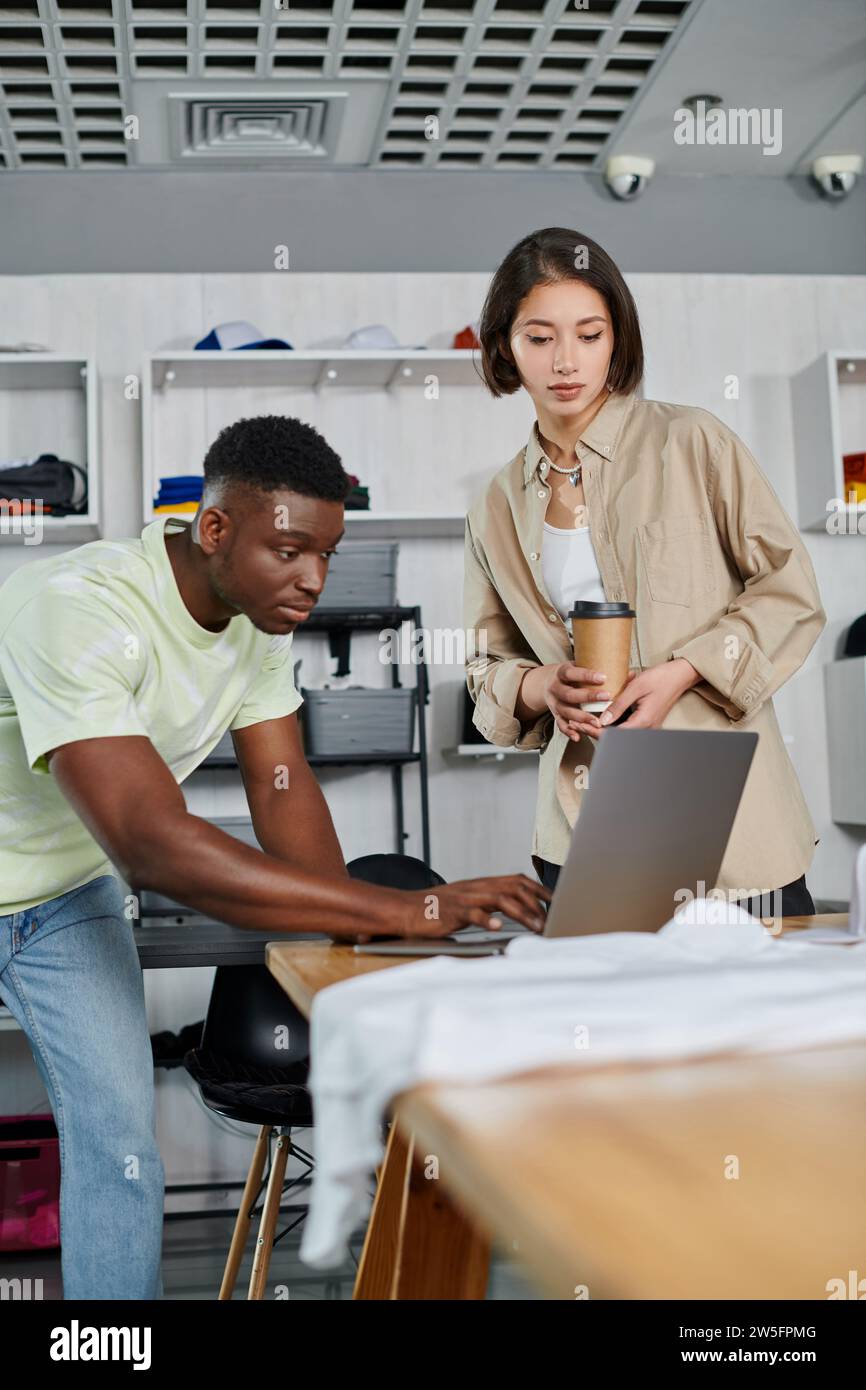 Inspirierter afroamerikanischer Mann mit Laptop in der Nähe asiatischer Frau mit Pappbecher im Bekleidungsdesign-Studio Stockfoto