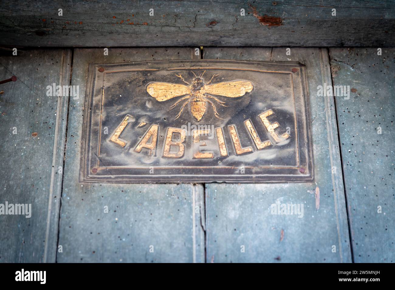 Ein altes Blechschild mit einer Biene und der französischen Schrift L'Abeille, was Biene bedeutet, auf einer dunklen Holzwand. Stockfoto