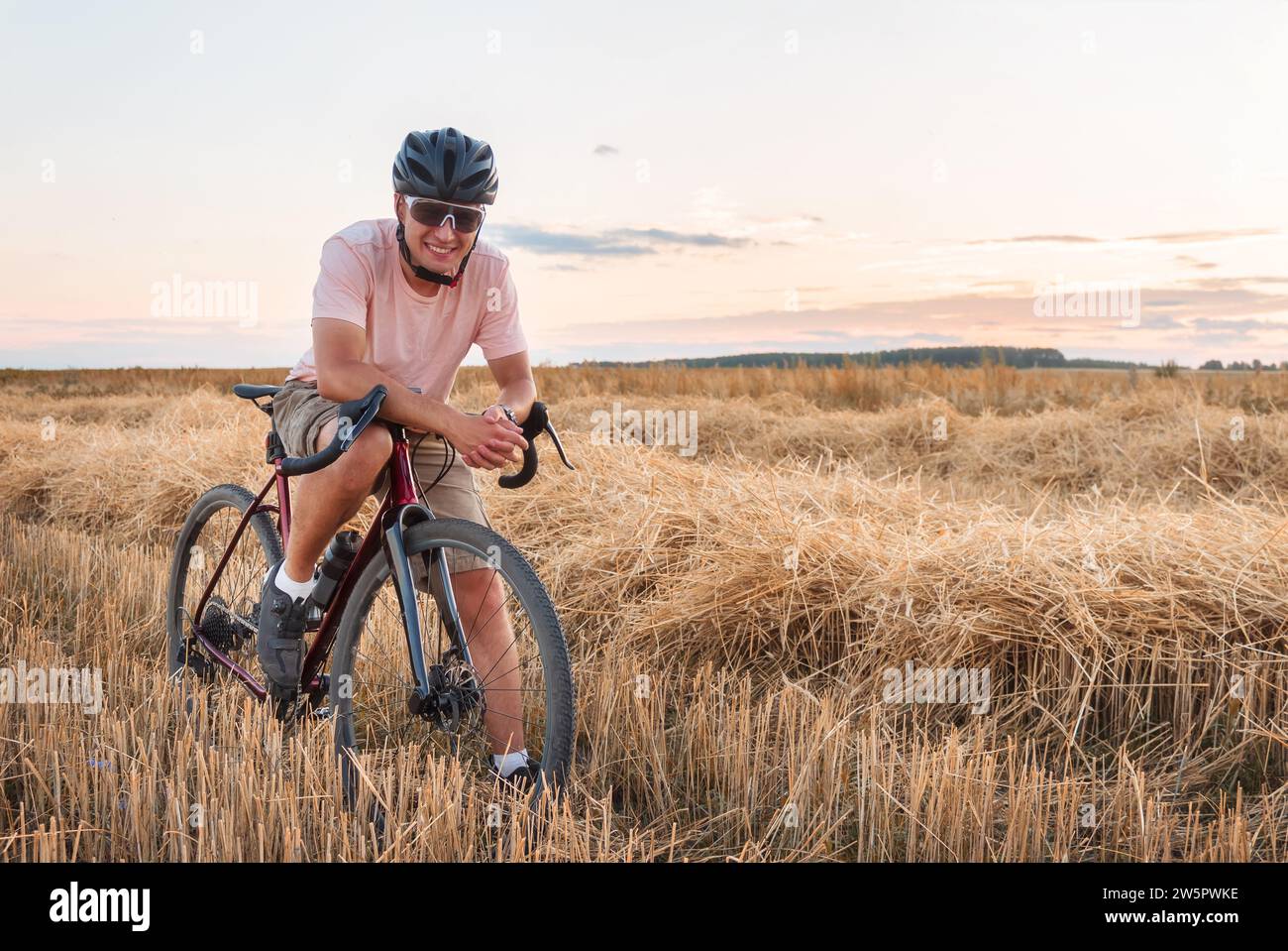 Radfahrer stehen bei Sonnenuntergang auf dem Feld. Wunderschöne Landschaft mit Mann in Helm und Sonnenbrille, Schotterfahrrad und trockenem Gras. Sportlicher Lifestyle. Stockfoto