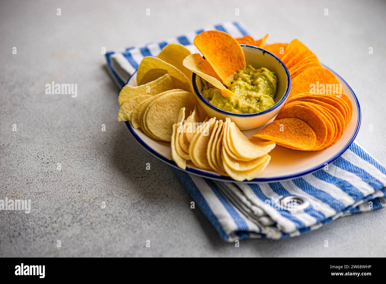 Fokusplatte mit verschiedenen Chips, einschließlich Kartoffeln, Paprika und Käse, serviert mit einer Schüssel mit frischer Avocado-Guacamole auf einer gestreiften Serviette Stockfoto