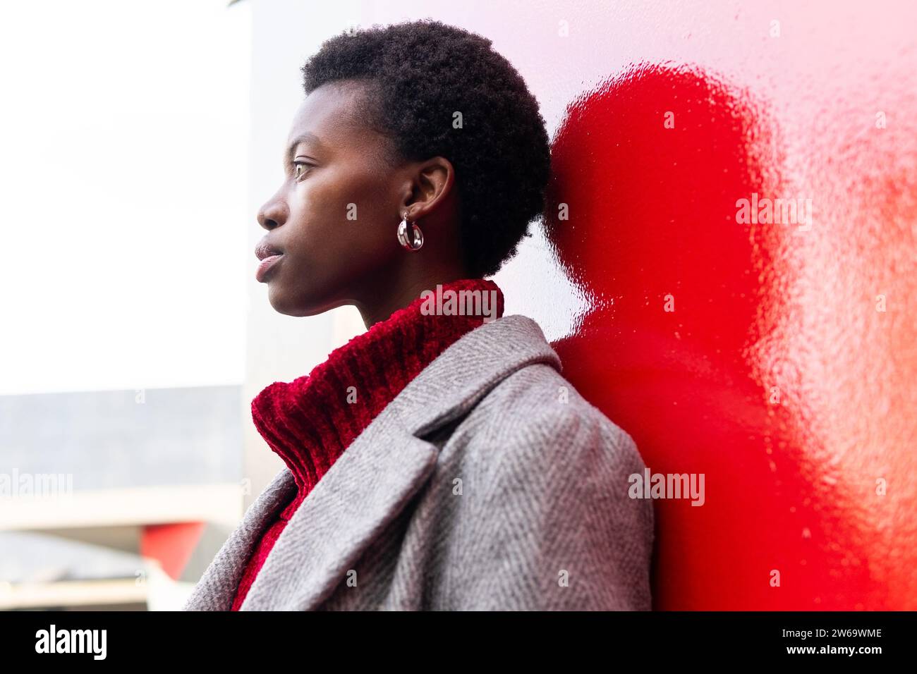 Seitenansicht einer schwarzen kurzhaarigen Geschäftsfrau in warmer grauer Jacke und Pullover, die sich an der roten Gebäudewand lehnt und im Sonnenlicht wegblickt Stockfoto