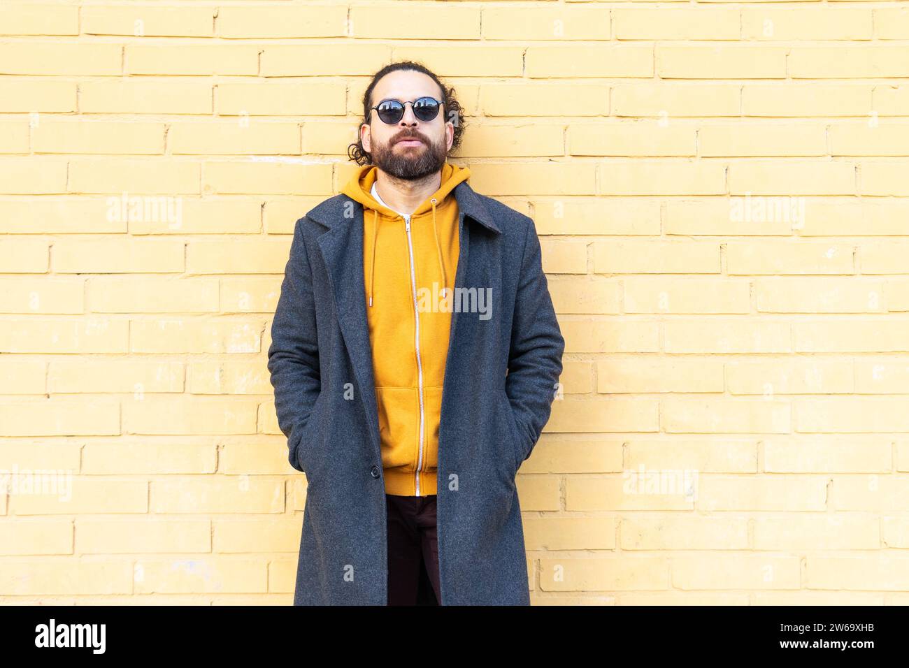 Ein entspannter Mann mit Sonnenbrille und gelbem Hoodie steht selbstbewusst vor einer gelben Ziegelwand und verkörpert urbanen Street Style. Stockfoto