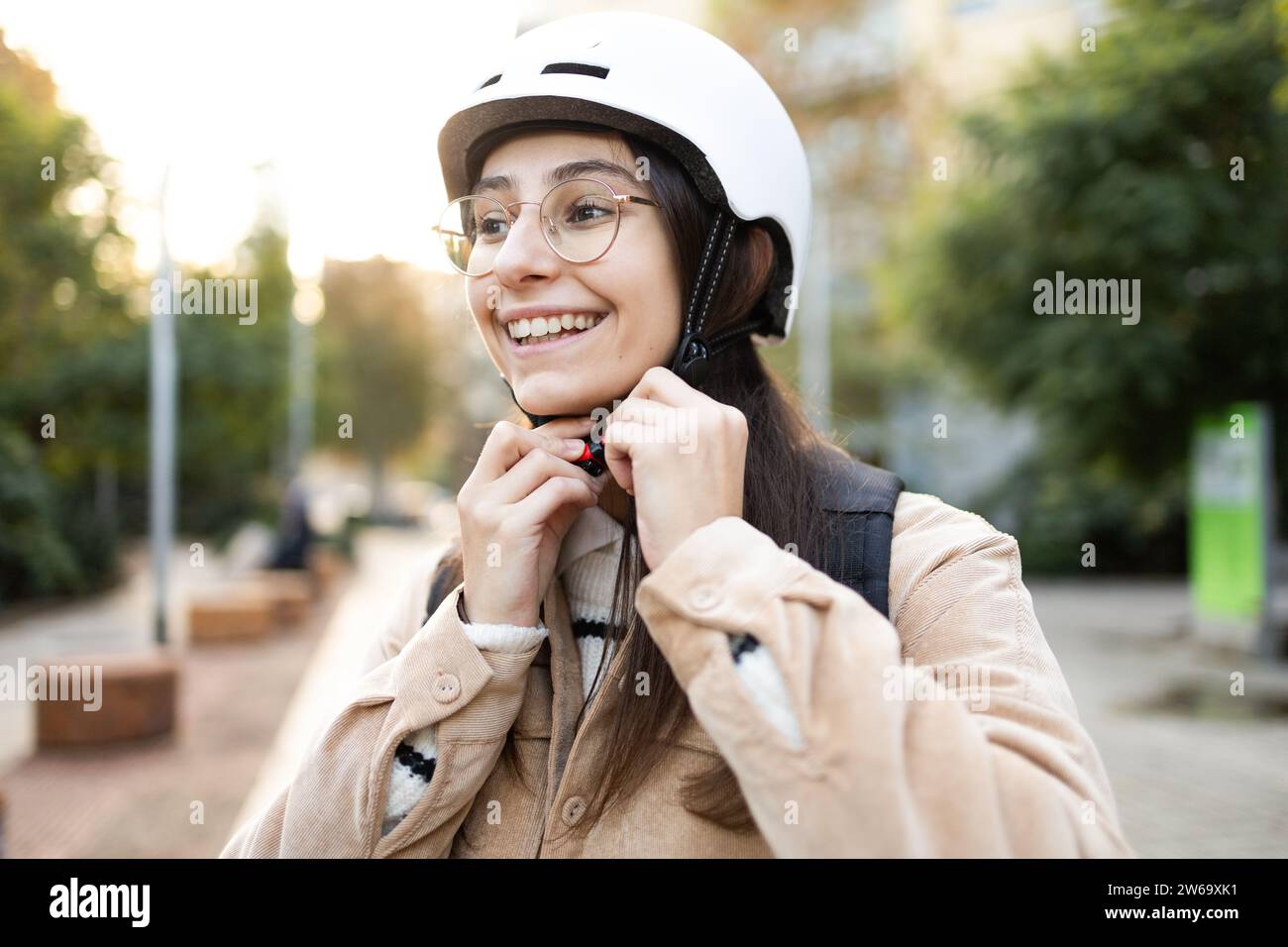 Eine fröhliche, junge Erwachsene Frau sichert den Riemen ihres weißen Helms im Freien und bereitet sich auf eine sichere Fahrt vor. Stockfoto