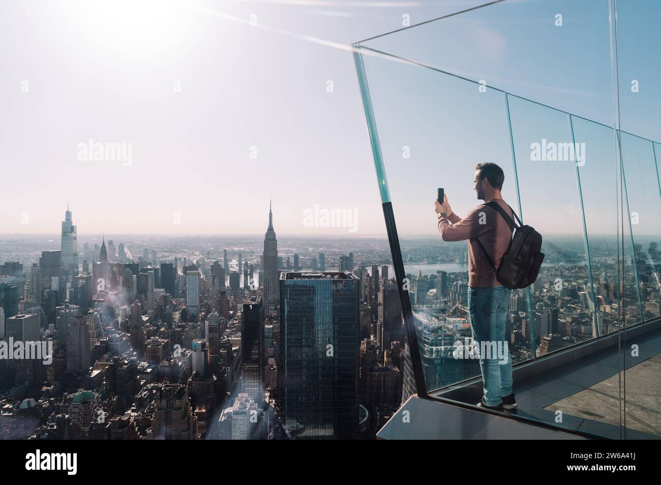 Ein Mann in lässiger Kleidung mit Rucksack macht mit seinem Smartphone ein Foto der weitläufigen Skyline von Manhattan, während die Stadt in sanftem Sonnenlicht getaucht ist. Stockfoto
