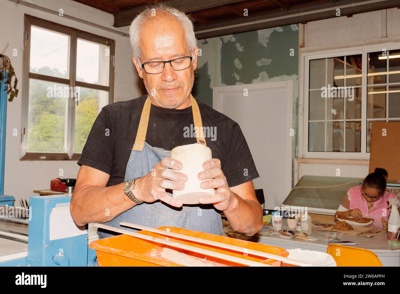 Portrait des älteren Handwerkers mit Schürze, der nach unten blickt, während er in der Keramikwerkstatt neben dem Tisch steht und zusätzliche Tonkanten mit einem Schaber auf cera auskratzt Stockfoto