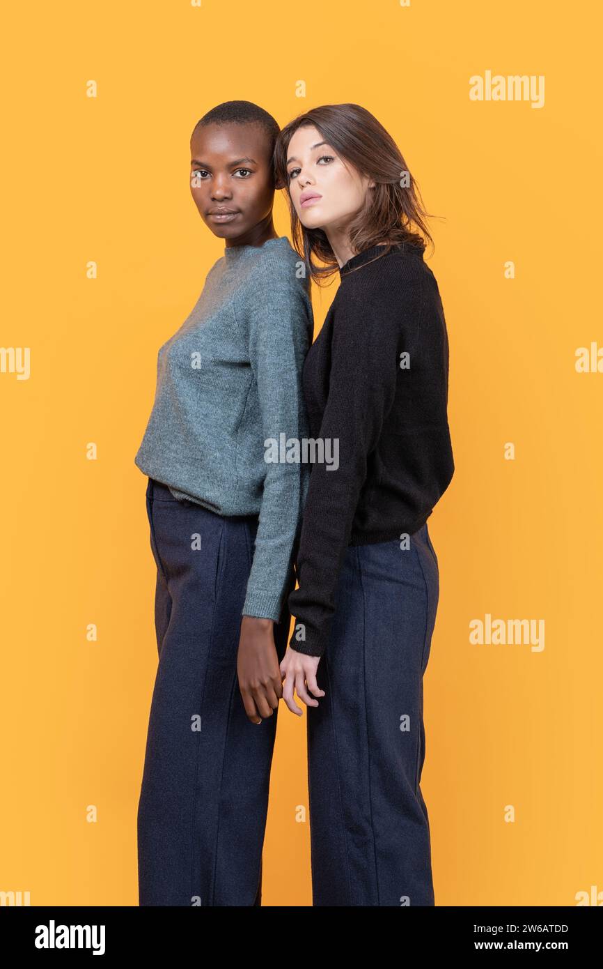 Seitenansicht des Mittelteils modischer multirassischer Frauen in lässiger Kleidung, die zusammen in die Kamera vor gelbem Hintergrund blicken Stockfoto