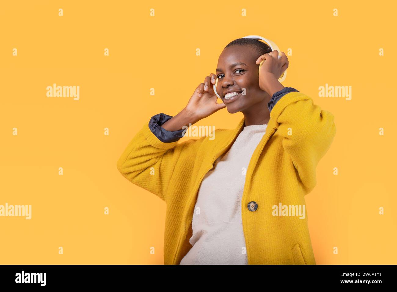 Fröhliche afroamerikanische junge Frau, die in die Kamera in der Jacke schaut und Musik über kabellose Kopfhörer vor gelbem Hintergrund hört Stockfoto