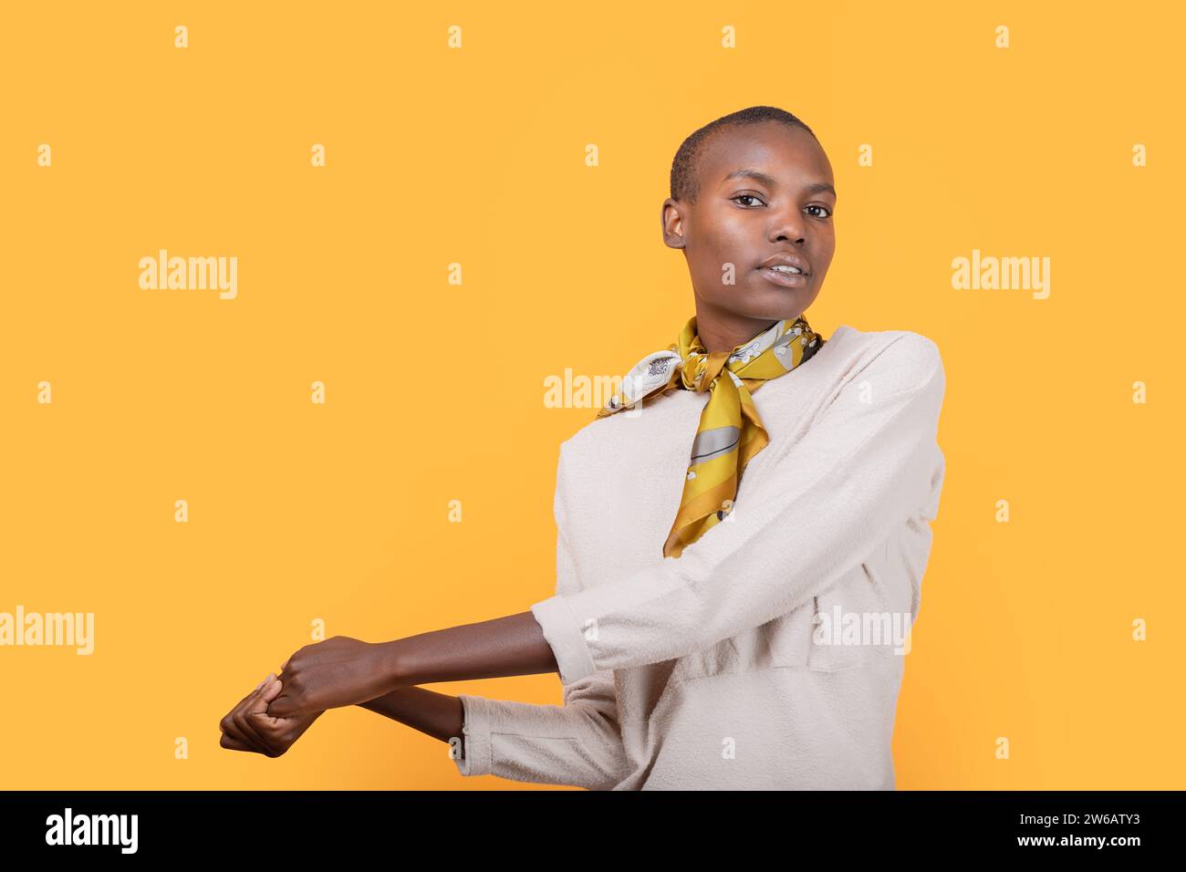 Selbstbewusste afroamerikanische schlanke junge Frau in modischer Freizeitkleidung, die einen Schal um den Hals trägt, während sie die Hände vor dem gelben Hintergras hält Stockfoto