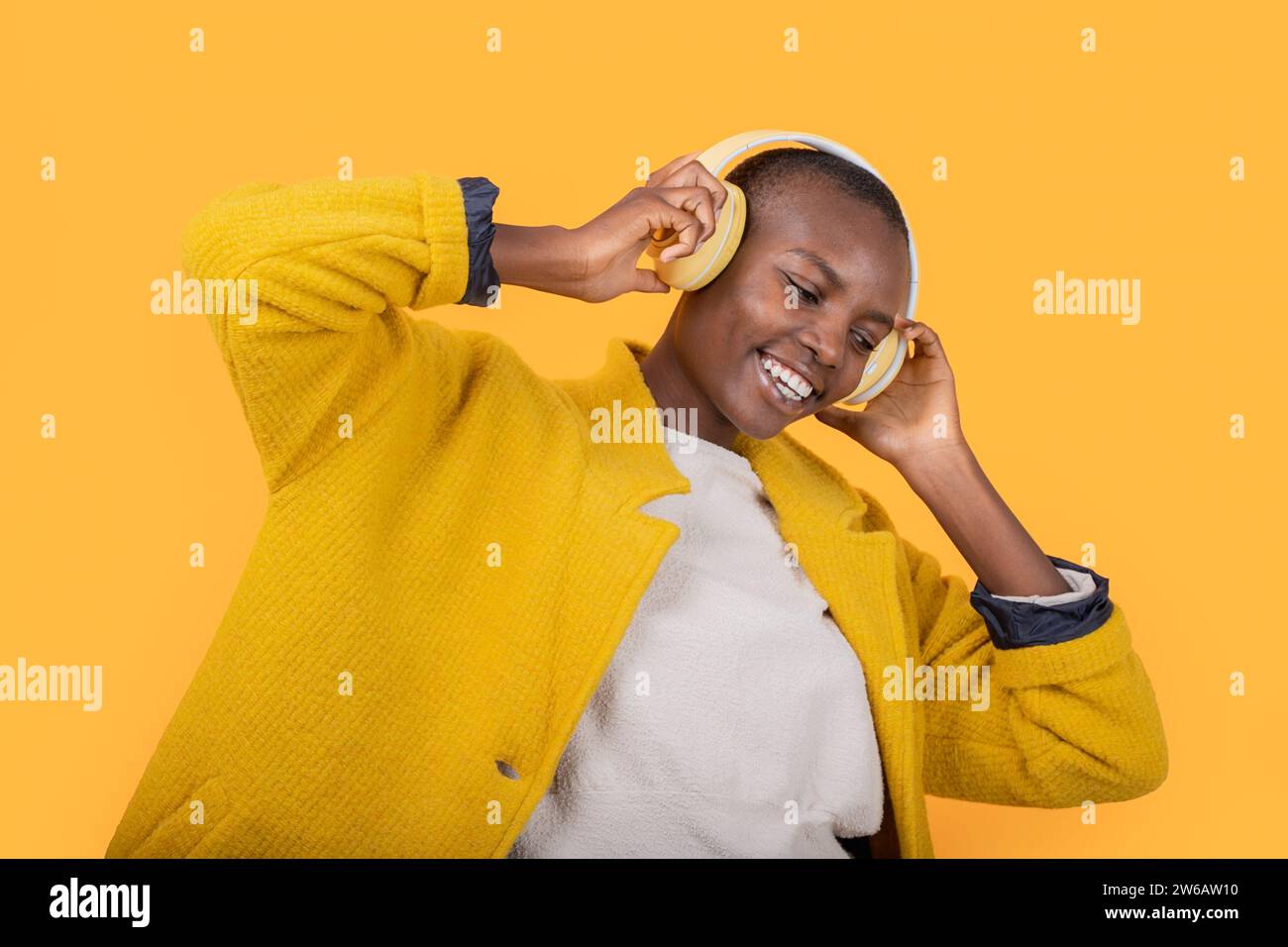 Fröhliche afroamerikanische junge Frau, die in der Jacke nach unten blickt und Musik über kabellose Kopfhörer vor gelbem Hintergrund hört Stockfoto