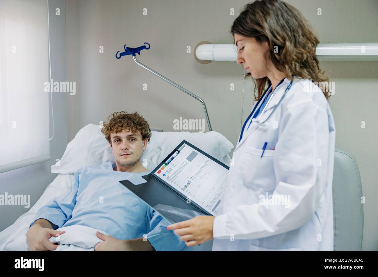 Seitenansicht einer jungen Ärztin mit Klemmbrett und Stethoskop in der Nähe des im Krankenhaus liegenden Patienten und beim Betrachten der Dokumente Stockfoto