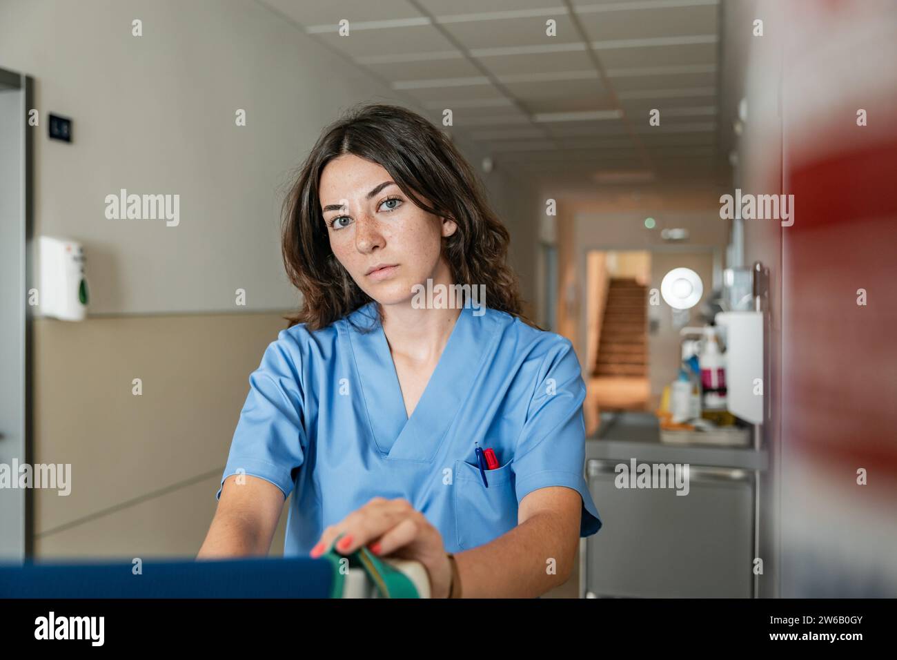 Junge Ärztin mit blauer Uniform und Blick in die Kamera, während sie im modernen Klinikkorridor vor verschwommenem Hintergrund sitzt Stockfoto