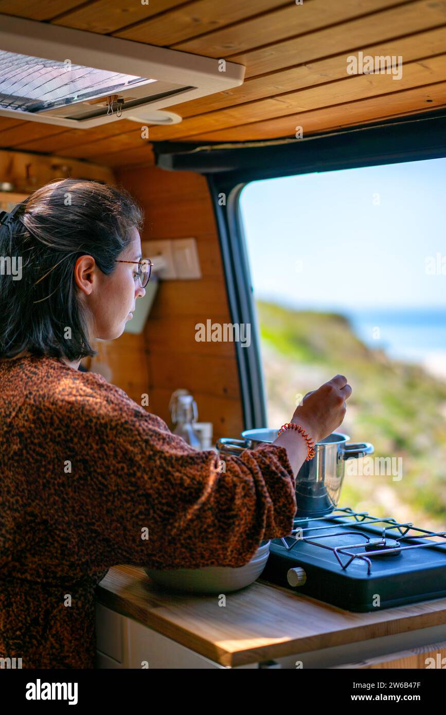 Seitenansicht einer fokussierten Reisenden in lässiger Kleidung und Brille, die während der Fahrt im Wohnmobil Essen auf einem Herd am Fenster in der modernen Küche kocht Stockfoto